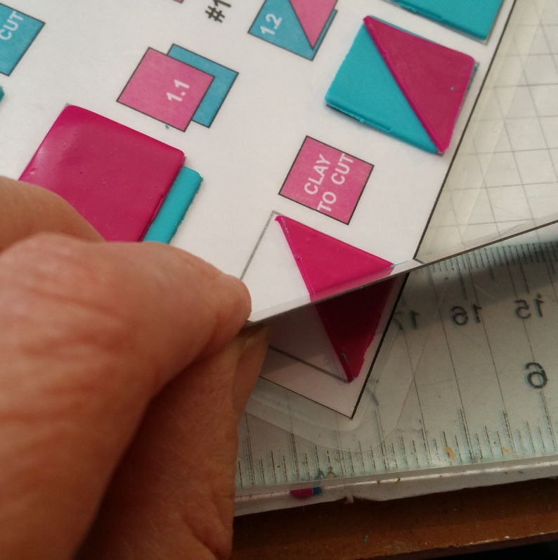 May include: A close-up of a hand holding a pink triangle of polymer clay against a white sheet of paper with a grid pattern. The paper has the words "Clay to Cut" printed on it.