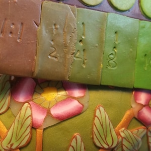 May include: Close-up of a set of polymer clay squares with numbers 12, 24, 18, and 1 written on them. The squares are arranged in a row and are different shades of green and brown.