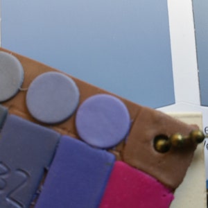 May include: A close-up of a paint color chart with a variety of blue and purple paint chips. The chart is made of brown cardboard and has a metal ring attached to it.
