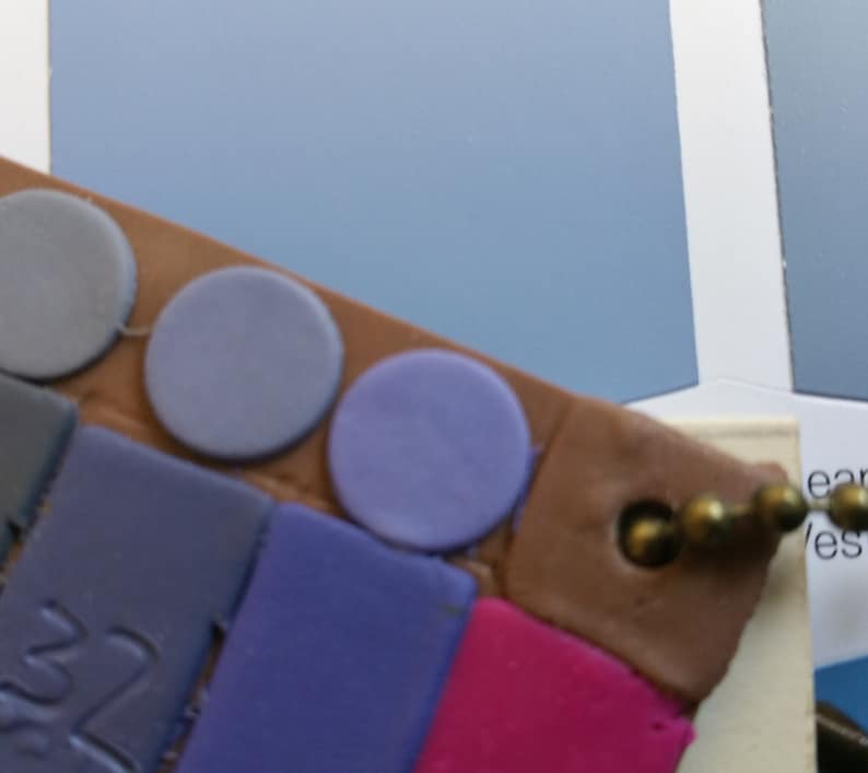 May include: A close-up of a paint color chart with a variety of blue and purple paint chips. The chart is made of brown cardboard and has a metal ring attached to it.