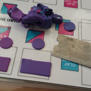 May include: Purple modeling clay on a white surface with a metal clay tool and a printed sheet with the words "CLAY TO CUT" and "CLAY CUT".