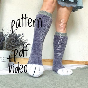 以下が含まれることがあります： 白い肉球が付いた、膝丈のグレーの靴下。靴下はふわふわした質感で、グレーとグリーンのリブ編みの袖口が付いています。画像には「pattern + pdf video」と「@happyknitties」と書かれています。