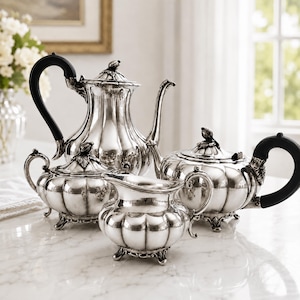 May include: A silver tea service set consisting of a tall teapot, a sugar bowl, a creamer, and a smaller teapot. Each piece features a fluted body, ornate leaf-shaped finials on the lids, and black curved handles with decorative metal accents.