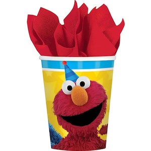 May include: A party cup featuring Elmo from Sesame Street, with a blue and white striped rim and a yellow background. The cup is filled with red tissue paper. Elmo is wearing a blue party hat.