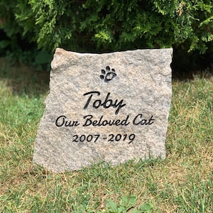May include: A gray granite stone engraved with a paw print, the name "Toby", the text "Our Beloved Cat", and the dates "2007-2019".