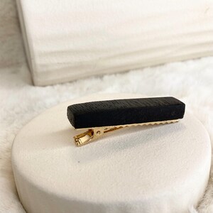 Könnte beinhalten: Eine schwarze rechteckige Haarklammer mit einem goldenen Metallverschluss. Die Klammer ist aus Holz und hat eine strukturierte Oberfläche.