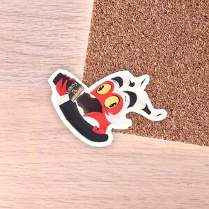 May include: A colorful sticker featuring a cartoon character with red, black, and white details. The character is holding a bottle of Guinness. The sticker is on a wooden surface.