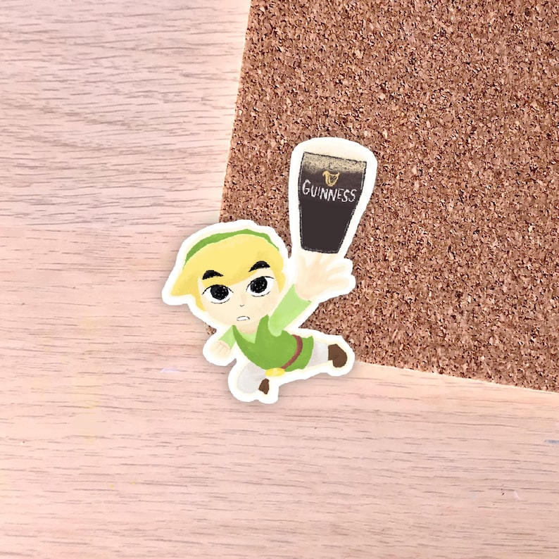May include: A sticker featuring a cartoon character in a green tunic and hat, holding a pint of Guinness. The Guinness glass is black with white foam and the Guinness logo. The sticker is on a light wood surface.