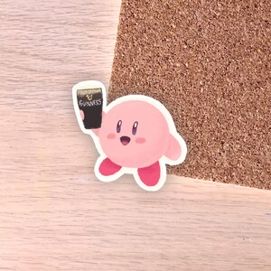 May include: A pink cartoon character sticker holding a pint of Guinness. The character has a round body, small arms and legs, and a smiling face. The Guinness pint is black with the brand name printed on it.
