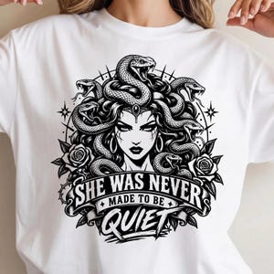 Peut inclure: Un t-shirt blanc avec une impression graphique noire représentant une femme aux cheveux en forme de serpents, entourée de roses et d'étoiles. Sous l'illustration, une bannière affiche le texte "SHE WAS NEVER MADE TO BE QUIET".