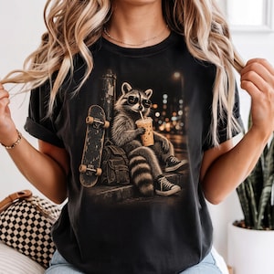 Puede incluir: Una camiseta negra con el gráfico de un mapache con gafas de sol y zapatillas, sentado en un bordillo mientras sostiene una bebida. Un monopatín se apoya contra un poste junto a una mochila, con un fondo de calle urbana por la noche.