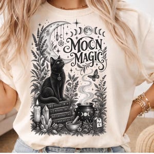 Peut inclure: Un t-shirt crème avec une illustration à l'encre noire représentant un chat assis sur une pile de livres intitulés Spells, Potions, Crystals et Moon Rituals. Le motif inclut un croissant de lune, un chaudron fumant, une bougie et le texte Moon Magic.