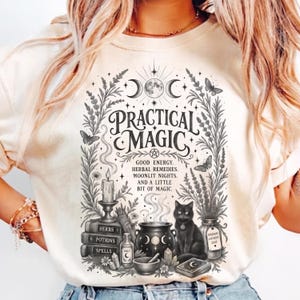 以下が含まれることがあります： 「PRACTICAL MAGIC」および「GOOD ENERGY, HERBAL REMEDIES, MOONLIT NIGHTS, AND A LITTLE BIT OF MAGIC」というテキストが黒いグラフィックでプリントされたクリーム色のTシャツ。デザインには黒猫、大釜、本、ハーブが描かれています。