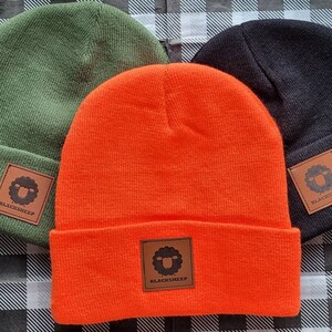 May include: Three knit beanies in different colors: green, orange, and black. Each beanie has a brown leather patch with a black sheep logo and the text "BLACKSHEEP".