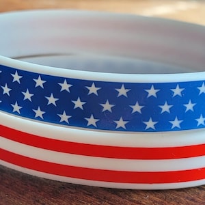 May include: Two white silicone bracelets with an American flag design. The top bracelet has a blue field with white stars and the bottom bracelet has red and white stripes.