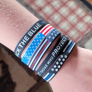 May include: Four black silicone wristbands with different designs. The top band says "Back the Blue" in white lettering. The second band has an American flag design. The third band says "Support Those Who Protect" in white lettering. The bottom band has a thin blue line with white stars.