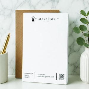 May include: White notepad with the text "ALEXANDER SMITH REALTY" in black. The notepad has a crown logo and contact information at the bottom. A gold pen rests in a white ceramic cup to the left.