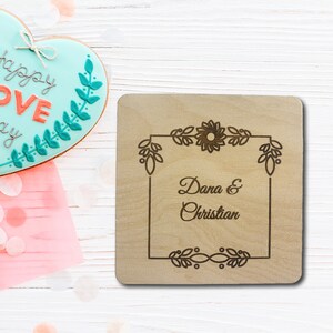 May include: A wooden square sign with a floral design and the text "Dana & Christian" engraved in the center.