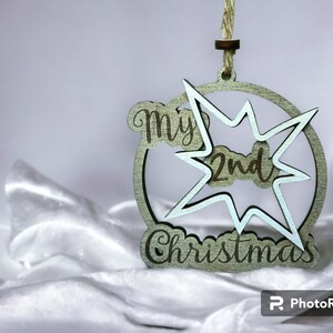 May include: A wooden Christmas ornament with the text "My 2nd Christmas" in a circle and starburst design. The ornament is light green with a natural twine hanger. The background is a white, silky fabric.