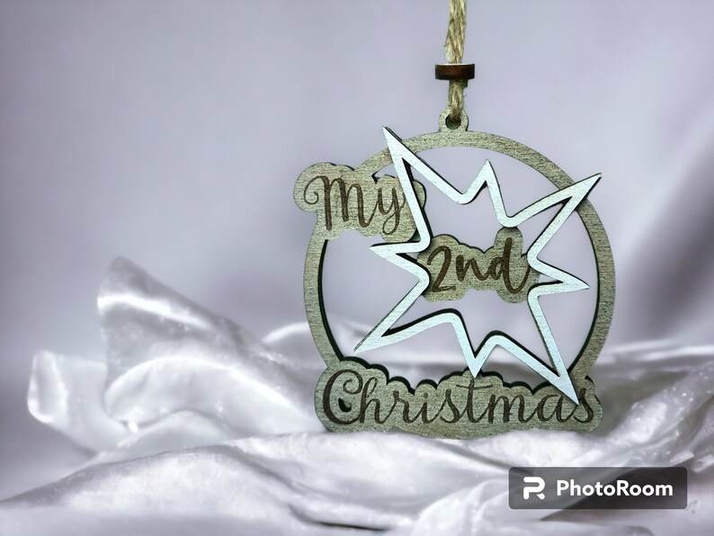 May include: A wooden Christmas ornament with the text "My 2nd Christmas" in a circle and starburst design. The ornament is light green with a natural twine hanger. The background is a white, silky fabric.
