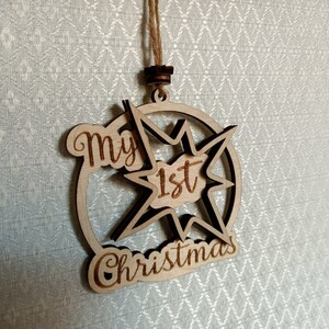 May include: Wooden Christmas ornament with the text "My 1st Christmas". The ornament is star-shaped with a circular border and a natural wood finish. It hangs from a twine string.