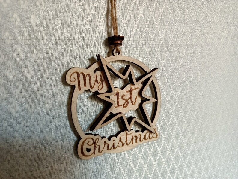 May include: Wooden Christmas ornament with the text "My 1st Christmas". The ornament is star-shaped with a circular border and a natural wood finish. It hangs from a twine string.