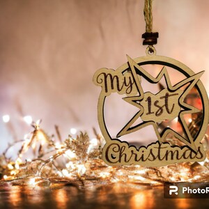 May include: Wooden Christmas ornament with the text "my 1st Christmas" in a starburst design. The ornament is made of light-colored wood and hangs from a natural twine string. The background features soft lighting and decorative elements.