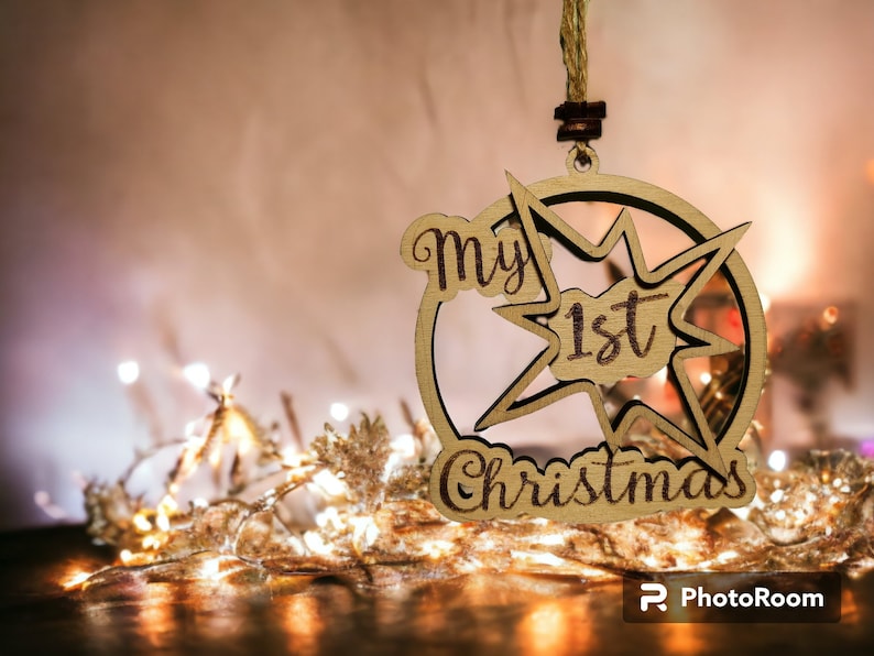 May include: Wooden Christmas ornament with the text "my 1st Christmas" in a starburst design. The ornament is made of light-colored wood and hangs from a natural twine string. The background features soft lighting and decorative elements.
