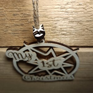 May include: Wooden Christmas ornament with the text "My 1st Christmas". The ornament is light brown with a starburst design and a small decorative element at the top. It hangs from a natural twine.