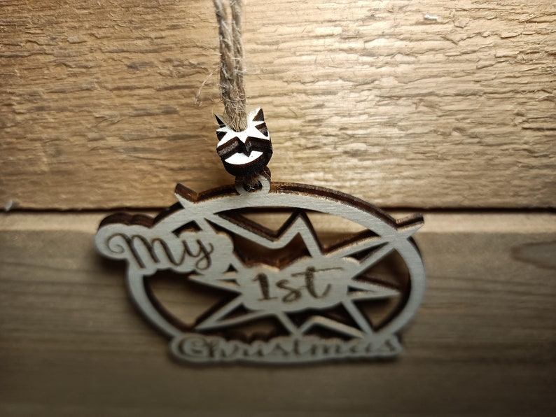 May include: Wooden Christmas ornament with the text "My 1st Christmas". The ornament is light brown with a starburst design and a small decorative element at the top. It hangs from a natural twine.