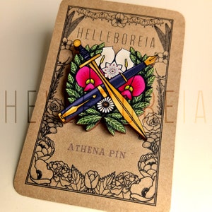 May include: Wooden Athena pin with a sword and pen design, set against a floral wreath with pink and white flowers. The pin is on a brown card with the text "HELLEBOREIA" and "ATHENA PIN".