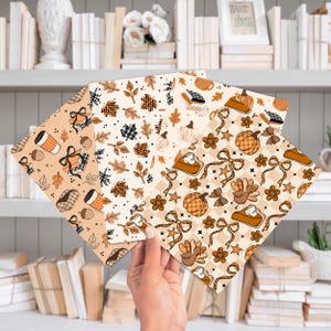 May include: A hand holds three sheets of patterned paper with fall-themed designs. The patterns include pumpkins, leaves, coffee cups, and pie slices in shades of brown, orange, and cream. The background shows a bookshelf.