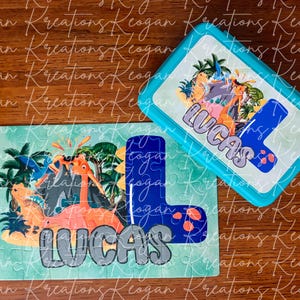 May include: A blue and green puzzle with a dinosaur theme. The puzzle is shaped like the letter L and has the name "Lucas" on it. The puzzle is next to a blue lunchbox with the same dinosaur theme and the name "Lucas" on it.