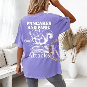 May include: A lilac-colored t-shirt with a graphic design on the back. The design features the text "PANCAKES AND PANIC" above a cartoon squirrel holding a fork over a stack of pancakes. The word "Attacks" is below the pancakes.