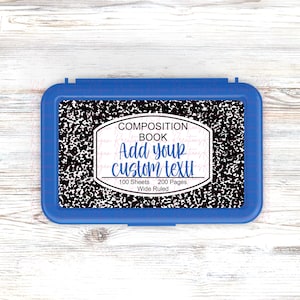 May include: A blue plastic pencil box with a composition book design. The composition book design has a black and white speckled pattern with the text "COMPOSITION BOOK Add your custom text! 100 Sheets 200 Pages Wide Ruled."