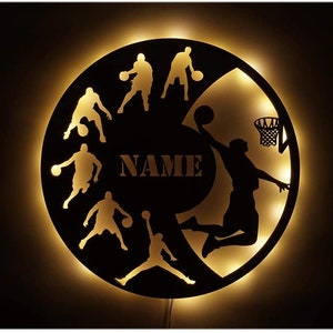 May include: A black and white silhouette of basketball players in various poses, including a player shooting a basket. The design is on a round, black, wooden plaque with a warm, yellow light shining through the cutouts. The word "NAME" is in the center of the plaque.