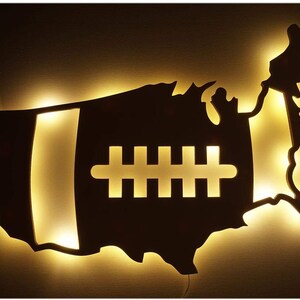 May include: A wooden cutout of the United States of America with a football shape in the center. The football has a white outline and laces. The cutout is illuminated from behind, creating a warm glow.