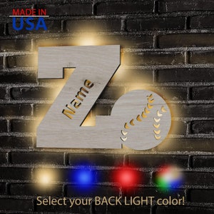 May include: Wooden letter Z night light with a baseball design. The letter is illuminated with warm white light and the baseball has a stitched design. The letter has the word "Name" cut out of it. The image shows four different colored light options: blue, red, green, and warm white.