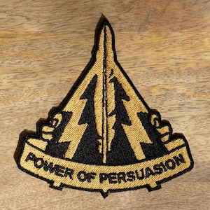 Puede incluir: Un parche bordado en oro y negro con un diseño triangular y el texto "POWER OF PERSUASION".