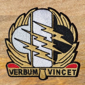 Può includere: Una toppa ricamata in oro con un designo di fulmine bianco e nero. La toppa ha una corona di alloro dorata che circonda i fulmini e le parole "Verbum Vincet" in lettere nere su uno stendardo rosso.