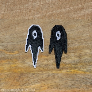 Op de afbeelding: Twee zwarte en witte geborduurde patches in de vorm van geesten met witte ogen. De patches bevinden zich op een houten oppervlak.