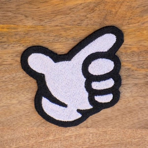 May include: White embroidered thumbs up patch with black outline. The patch is on a wooden surface.