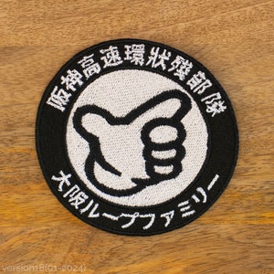 Peut inclure: Patch brodé noir et blanc avec du texte japonais qui se lit "Département d'intervention d'urgence à grande vitesse" et "Académie de surf Big Wave". Le patch présente une main de dessin animé blanche faisant un pouce en l'air.