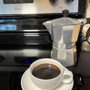 May include: A white ceramic coffee cup with a saucer, filled with dark coffee, sits on a black stovetop next to a silver and black stovetop espresso maker.