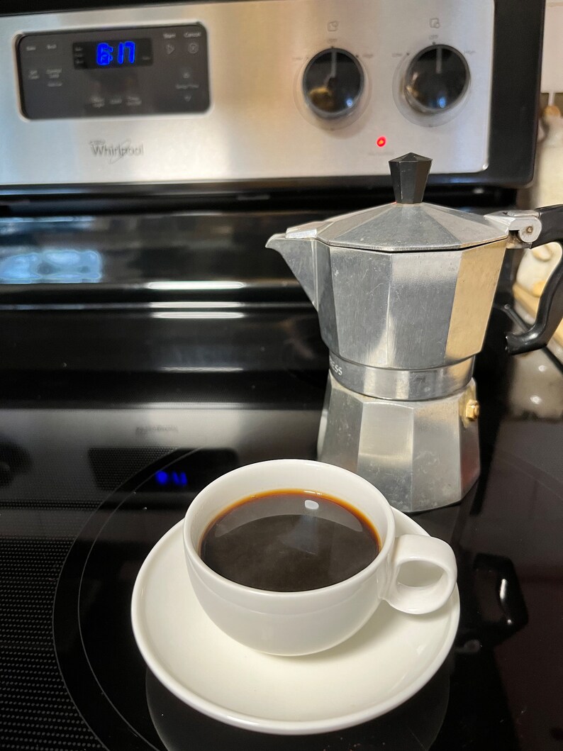 May include: A white ceramic coffee cup with a saucer, filled with dark coffee, sits on a black stovetop next to a silver and black stovetop espresso maker.