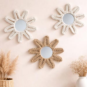 May include: Three daisy-shaped wall mirrors. Two are white, and one is tan. Each mirror has a circular reflective surface surrounded by woven, petal-like designs. The mirrors are mounted on a light pink wall, with decorative plants in the foreground.