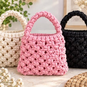 May include: Three small handbags crafted from thick cord in a macrame knot pattern. The bags are arranged in a row, featuring a cream-colored bag on the left, a bright pink bag in the center, and a solid black bag on the right side.