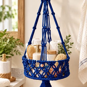 May include: A vibrant royal blue macrame hanging basket featuring a circular base with intricate knotted patterns, suspended by four thick cords from a wooden ring. The basket holds white towels, a soap dispenser, and a natural sponge.