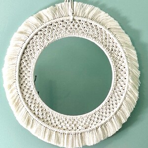 May include: Round macrame mirror with a cream-colored woven design and fringe. The mirror is centered within the macrame frame, creating a decorative wall hanging. The mirror is hanging on a light blue wall.