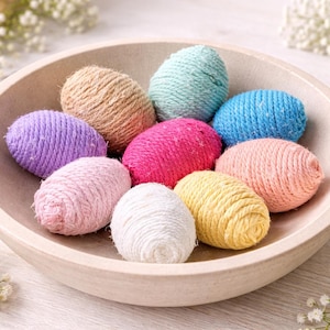 May include: A collection of colorful, yarn-wrapped Easter eggs in a light beige bowl. The eggs are in various colors, including pink, blue, yellow, and purple. The bowl is surrounded by white flowers, creating a festive spring theme.
